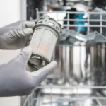 Une personne avec les mains gantées qui tient un filtre devant un lave-vaisselle ouvert