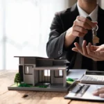 Un homme qui tend une clé à une autre personne devant une maquette de maison